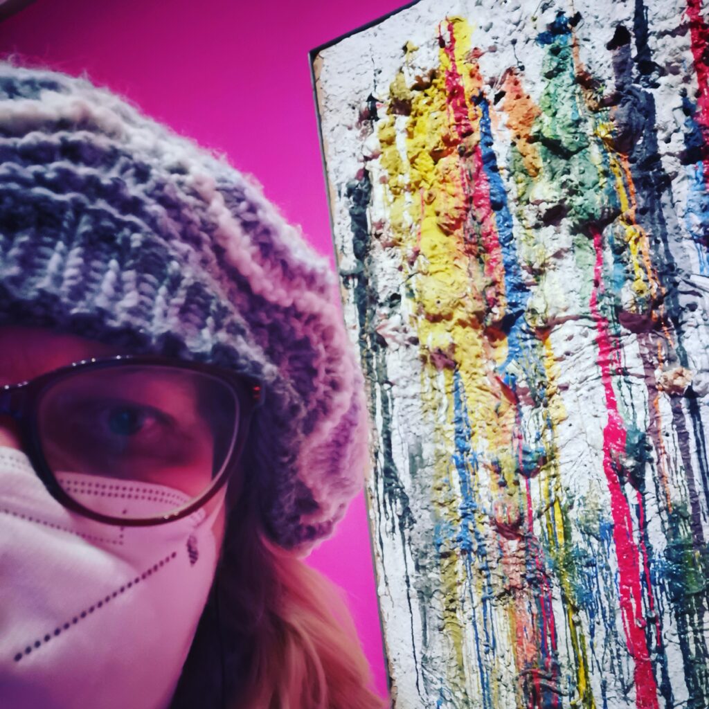 Bildbeschreibung: Claudia mit Mütze, Brille und Maske nur halb im Bild, im Hintergrund ein Ausschnitt aus einem Bild von Niki de Saint Phalle
