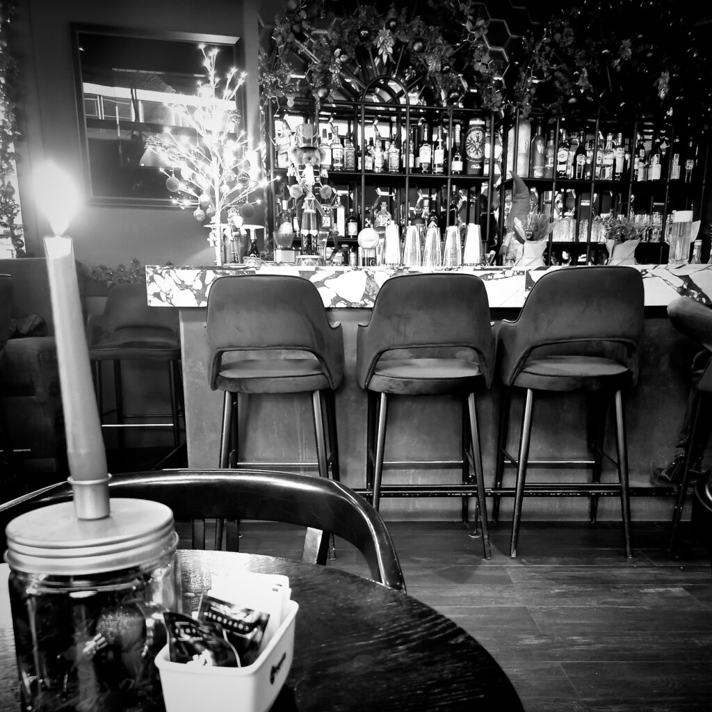 Foto in schwarzweiß, Innenraum eines Cafés mit Theke, Barhocker und Weihnachtsdeko. Im Vordergrund eine brennende Kerze auf einem Tisch.  