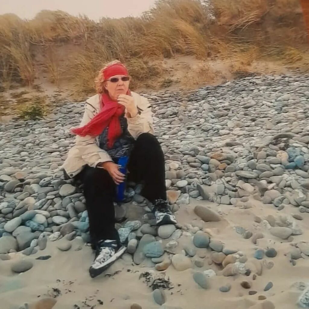 Claudia sitzt mit heller Regenjacke und einem roten Schal an einem Strand mit großen Steinen. Sie isst etwas.